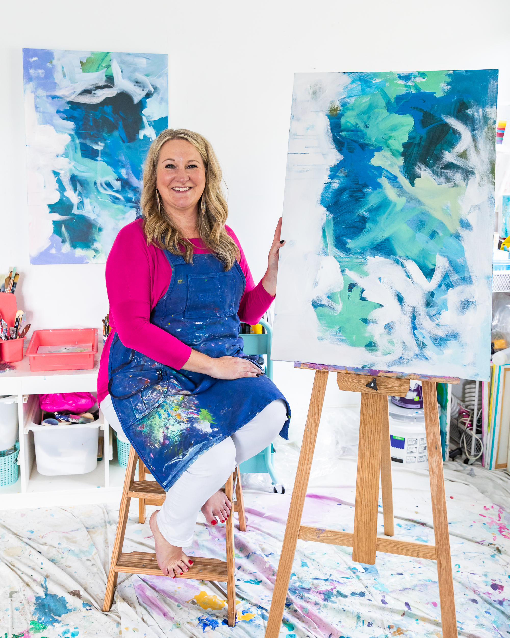 Heather Eck is an artist in her studio sitting on a bench next to a canvas with a blue and white painting.